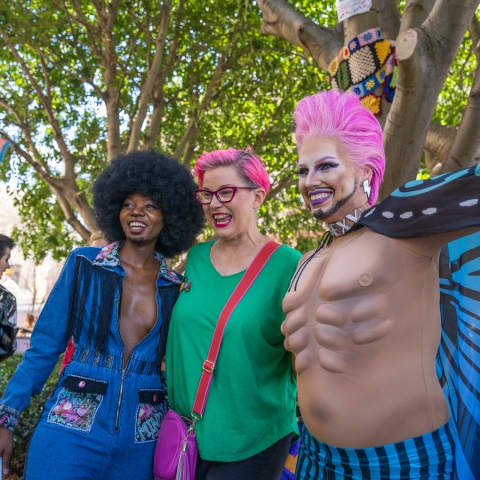 A group of three people wearing brightly coloured outfits. One person with pink hair, a bare chest and blue butterfly wings, another with pink hair, glasses and bag plus a bright green tshirt, and another with tasselled denim overalls and a large black afro